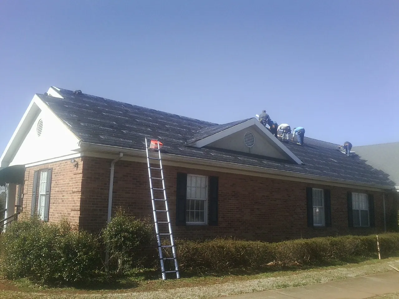 Crew tearing shingles off a brick ranch roof with a ladder set — Metal Roof Repair work by our crew in Lebeau