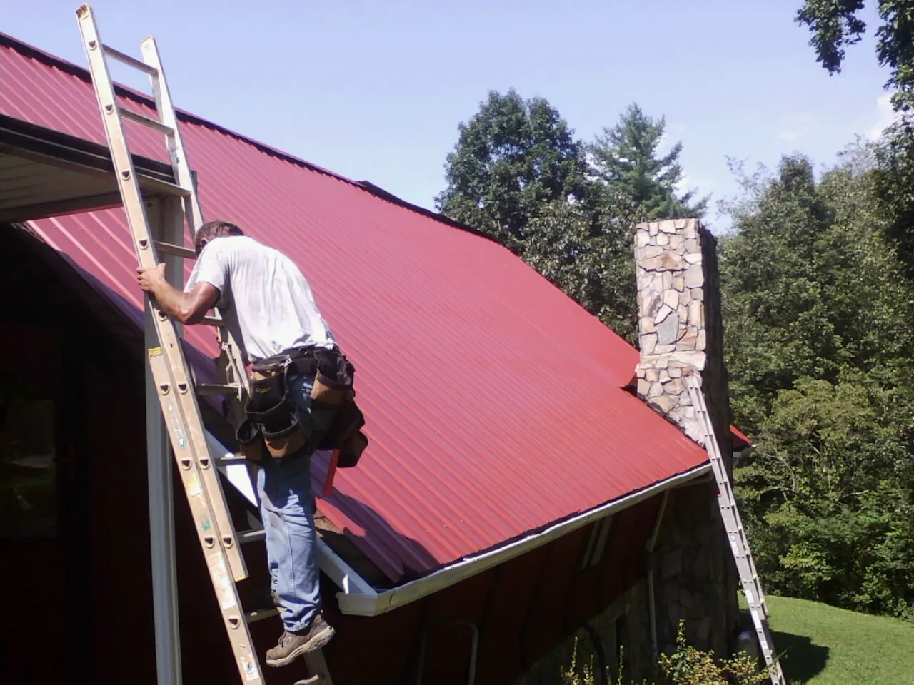 Roofer on a ladder beside a red metal roof and stone chimney — Soffit & Fascia Repair by Roofing Brothers in Lebeau